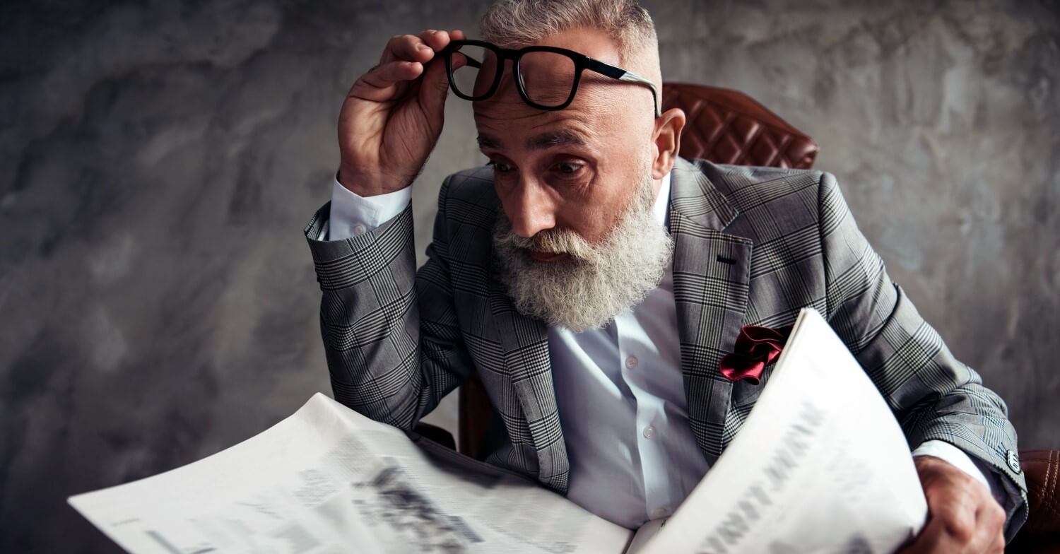 Ein Rentner mit grauem Bart studiert die Aktienkurse in der Zeitung, die Brille hat er dabei an die Stirn gehoben