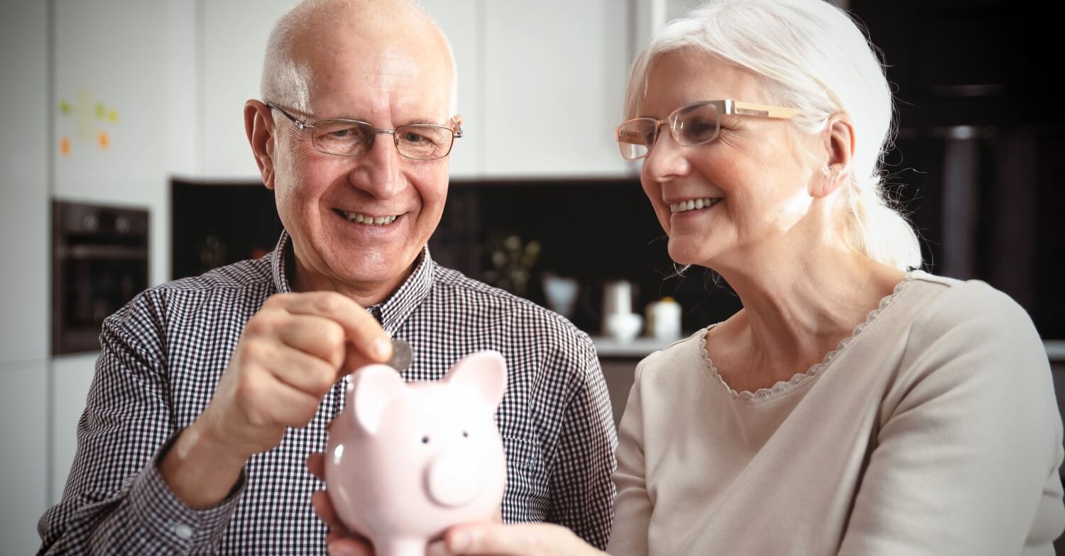 Ein Rentnerpaar hält ein Sparschwein in der Hand und füttert es mit Münzen. Das Paar sieht dabei glücklich aus