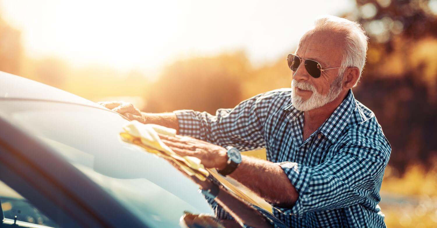 Ein grauhaariger Senior mit Sonnenbrille poliert die Windschutzscheibe seines Autos