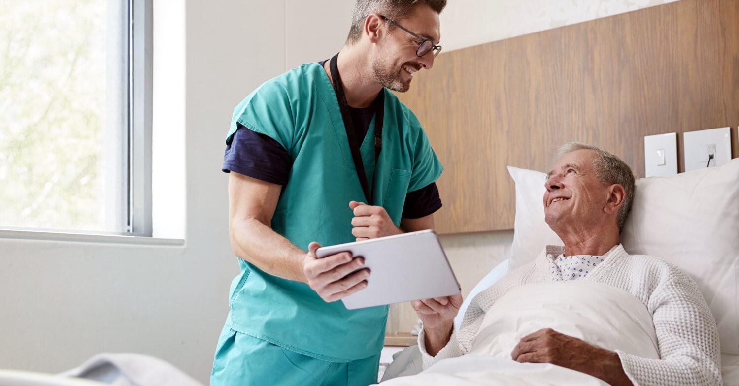Ein Pfleger im grünen Kittel steht bei einem Patienten im Krankenhausbett und unterhält sich mit ihm