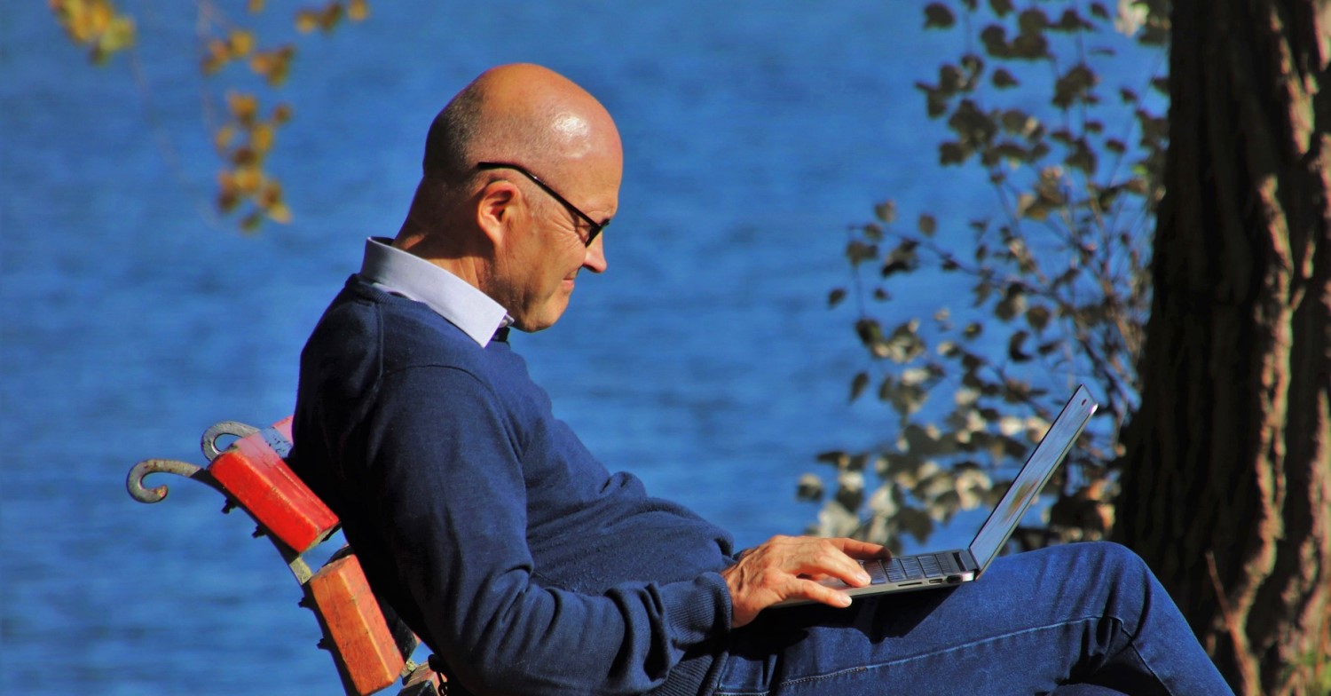 Ein älterer Mann mit Glatze und Brille sitzt auf einer Parkbank am Wasser und hat ein Notebook auf dem Schoß, auf dem er tippt