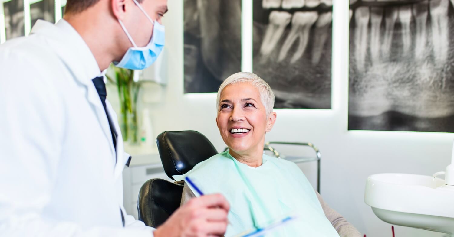 eine Seniorin sitzt im Behandlungsstuhl beim Zahnarzt. Der Zahnarzt trägt einen Mundschutz und ist der Patientin zugewandt, im Hintergrund hängen Röntgenaufnahmen ihres Kiefers