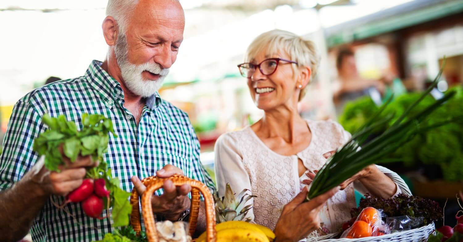 Ein Rentnerpaar beim Einkauf: beide haben Einkaufskörbe vor sich, die mit Obst und Gemüse gefüllt sind.