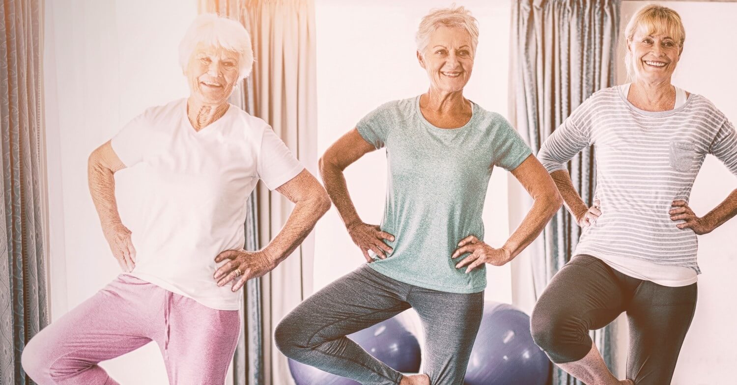 Drei Seniorinnen machen Yoga. Sie tragen Sportbekleidung und stehen jeweils auf einem Bein, das andere Bein ist angewinkelt, die Hände in die Hüften gestützt, der Oberkörper aufrecht
