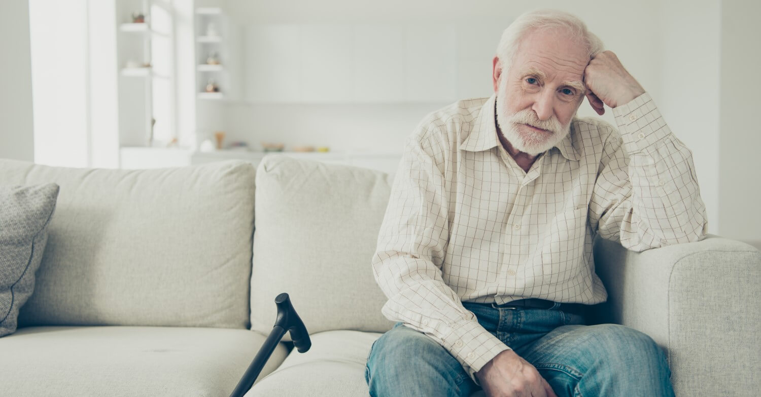 Ein Rentner sitzt alleine auf dem Sofa, neben ihm sein Gehstock. Er hat den Ellenbogen auf die Sofalehne gestützt und hält sich den Kopf.