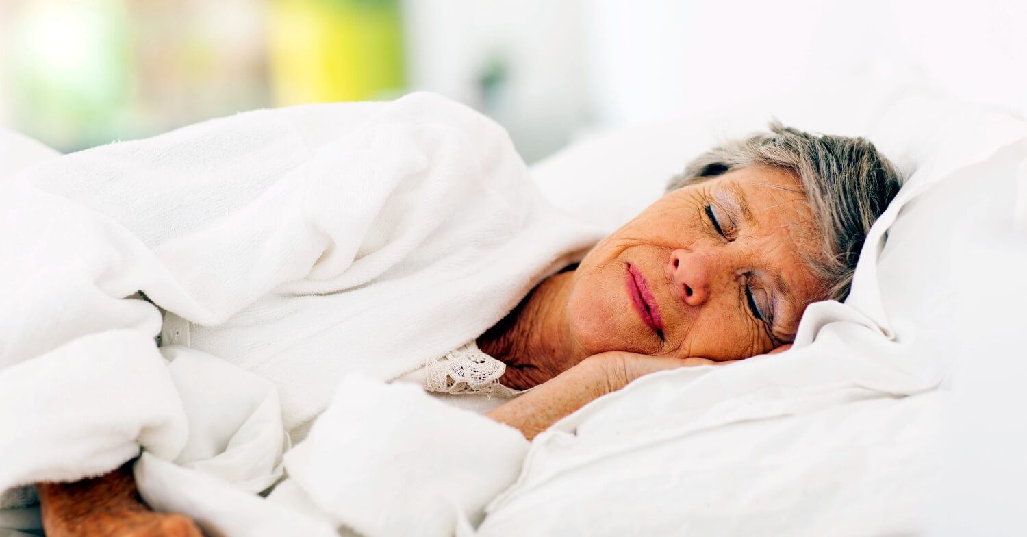 Eine Seniorin schläft in Seitenlage, sie hat die Hand unter den Kopf geschoben, der in Richtung Kamera blickt