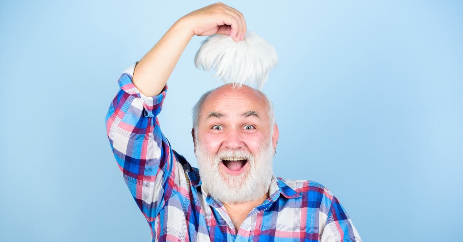 Ein Senior lacht in die Kamera, mit einer Hand hebt er seine Perücke hoch, darunter ist eine Glatze zu sehen