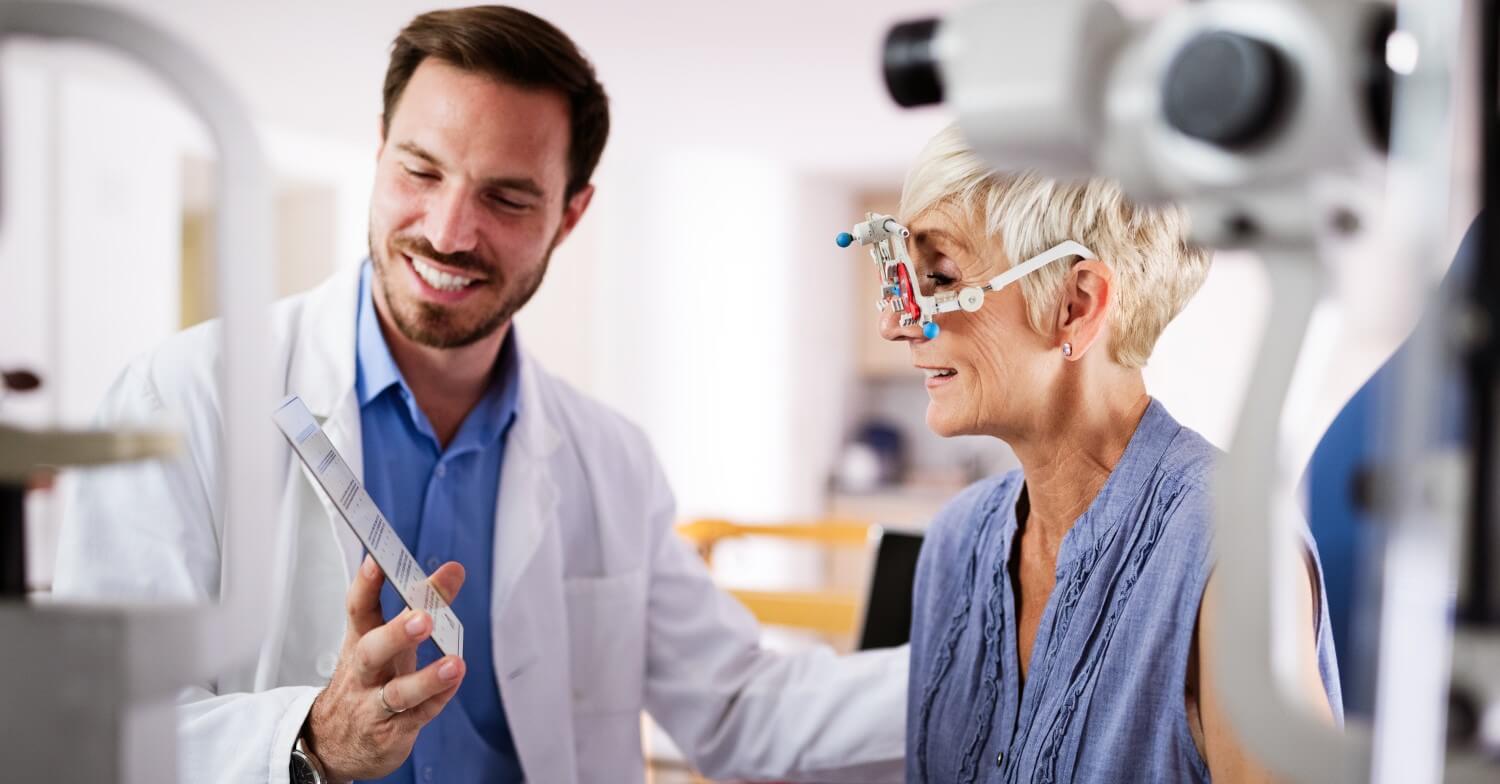 Eine grauhaarige Rentnerin ist beim Augenarzt und macht dort einen Sehtest. Der Augenarzt hält die Tafel mit den Buchstaben