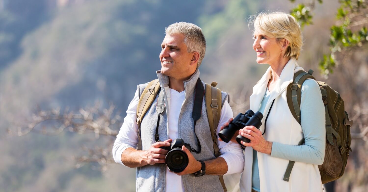 Rentnerpaar in der Natur, er hält einen Fotoapparat, sie ein Fernglas in der Hand