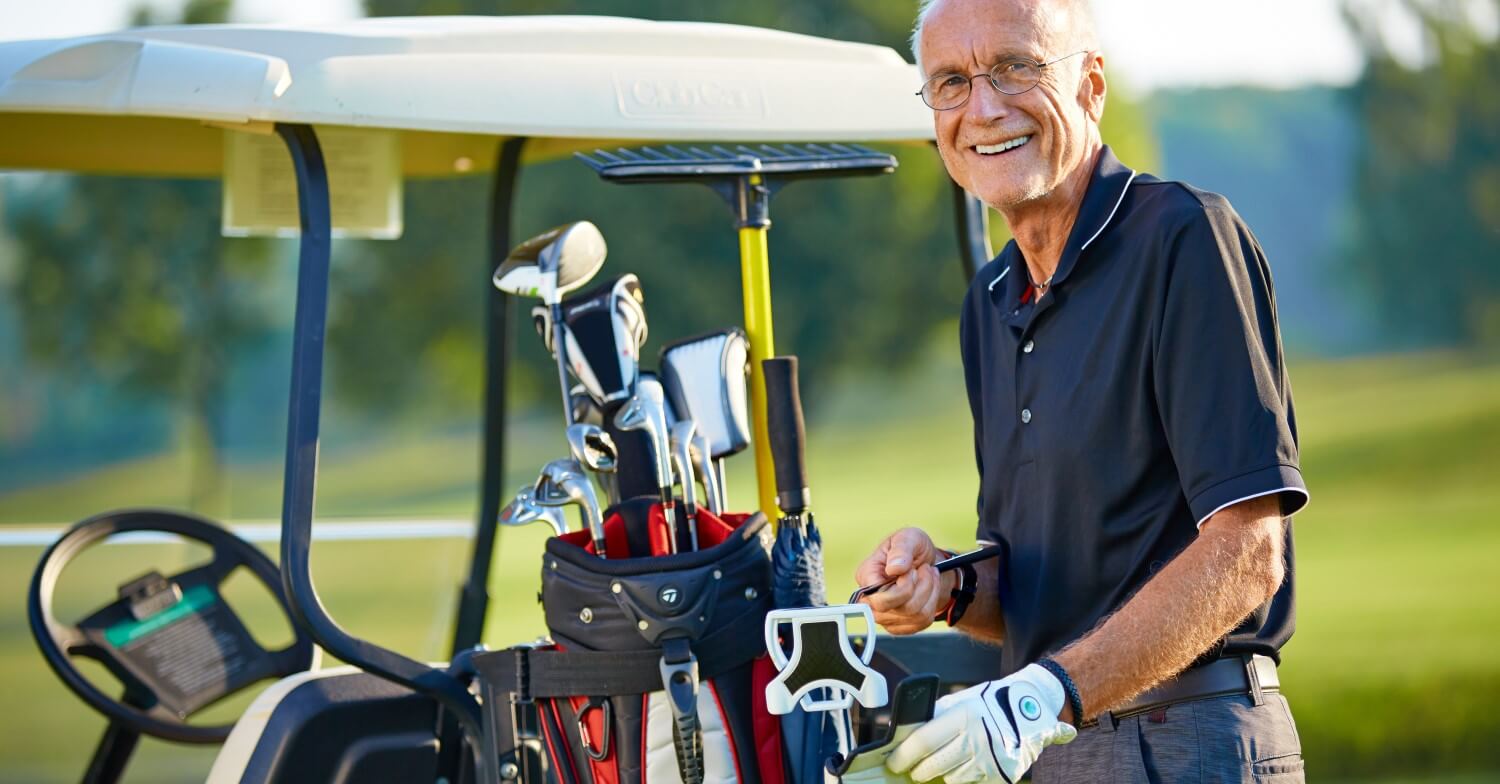 Ein Senior steht neben seinem Golfcart, er schaut in die Kamera und hantiert an der Tasche mit den Golfschlägern