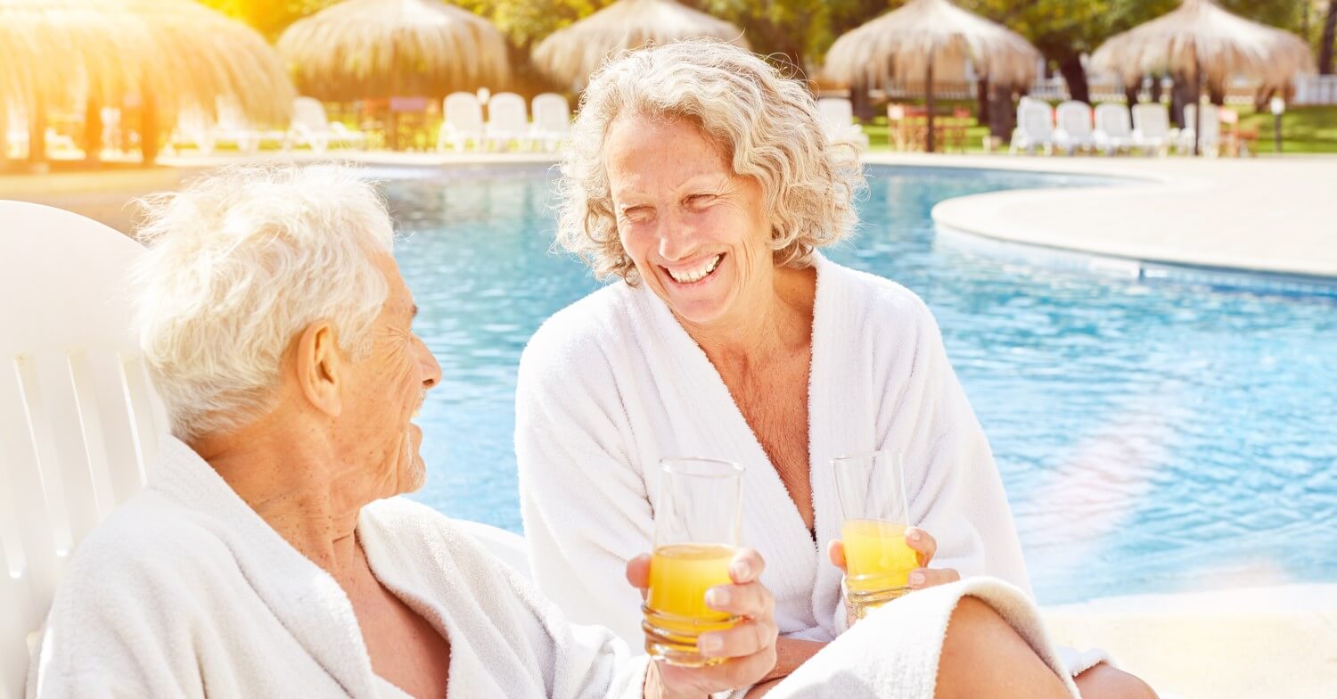 Zwei Senioren sitzen in Bademänteln am Pool und trinken Orangensaft