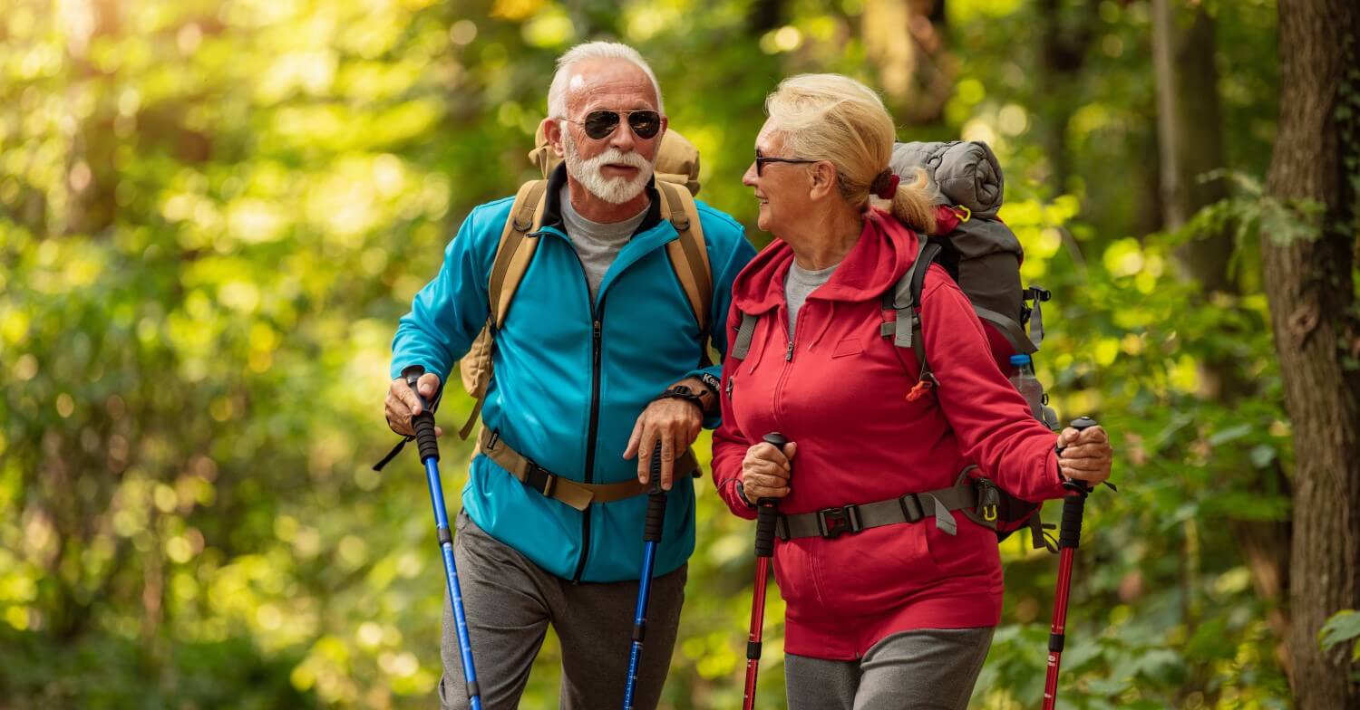 Ein Rentnerpaar bei einer Wanderung im Wald. Beide tragen Rucksäcke, Outdoorjacken und Wanderstöcke