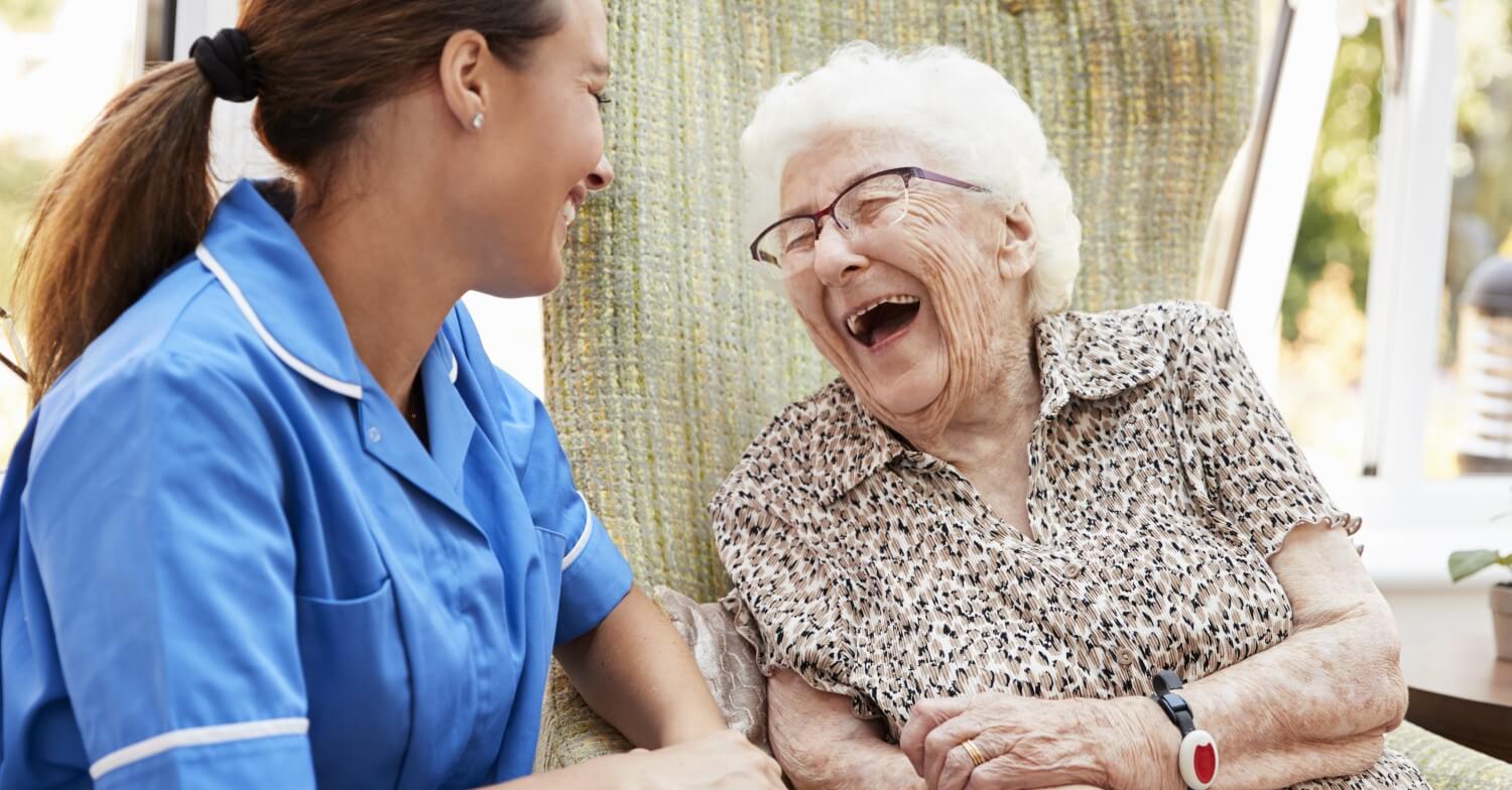 Eine Pflegerin in blauem Kittel unterhält sich mit einer Seniorin, die fröhlich lacht
