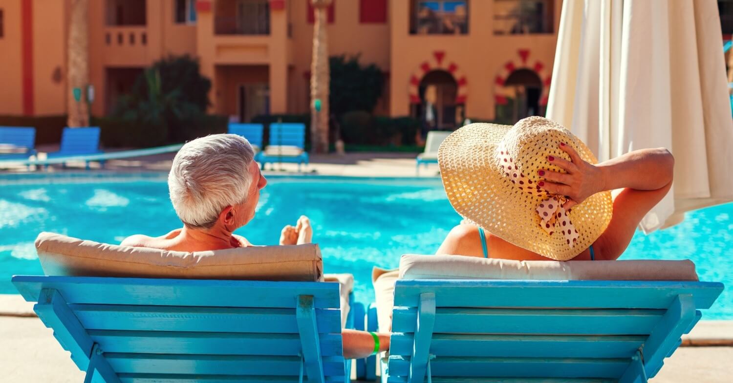 Ein Rentnerpaar sitzt auf blauen Liegestühlen am Pool und genießt die Sonne