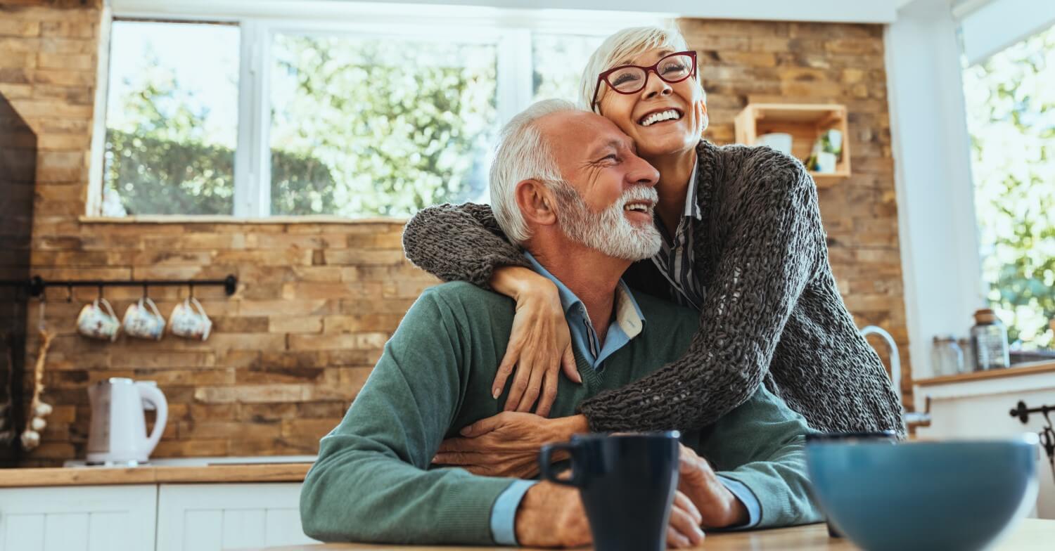 Ein Rentnerpaar sitzt in der Küche, umarmt sich und lacht dabei
