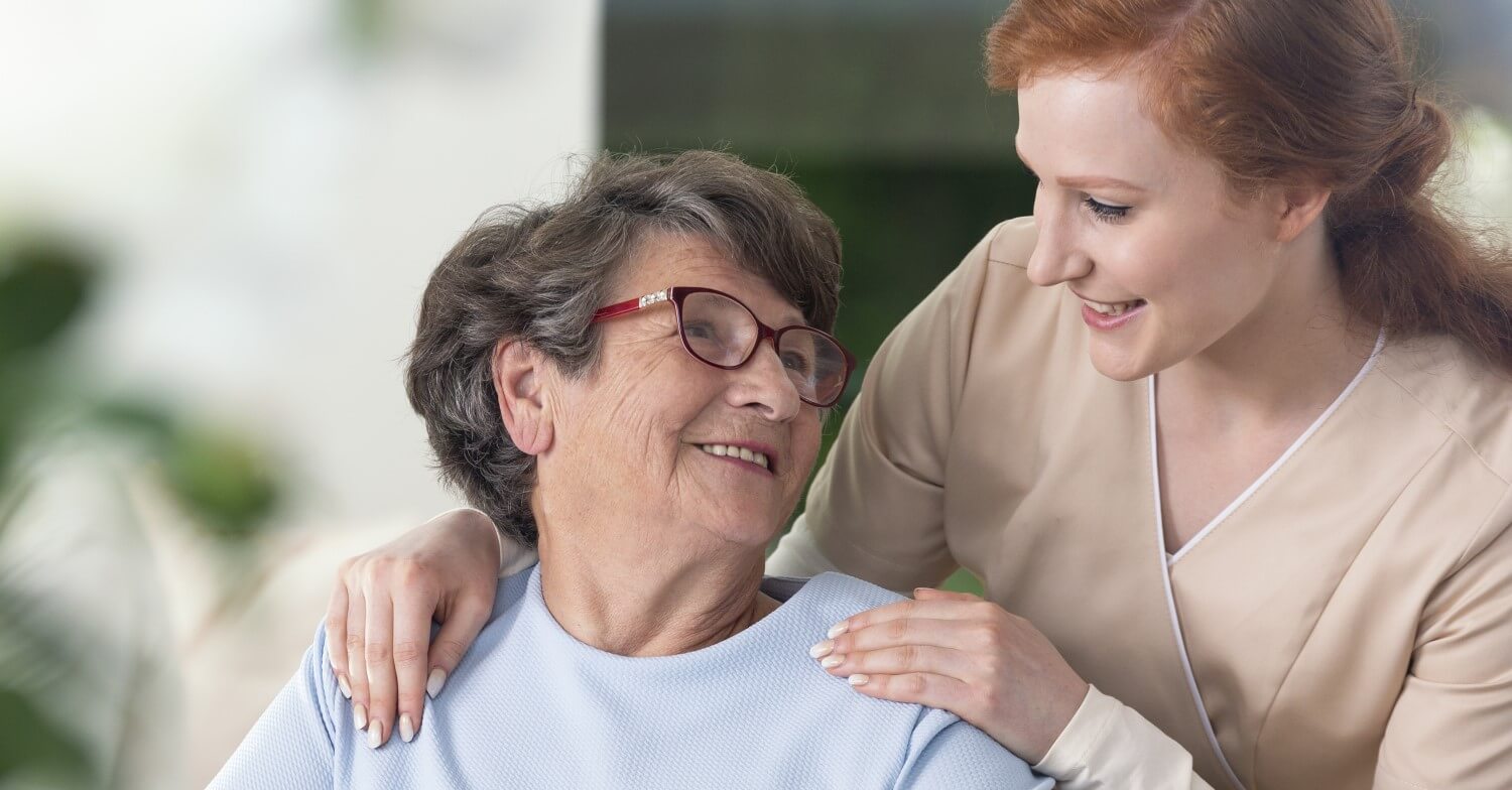 Eine Pflegerin mit roten Haaren kümmert sich um eine Seniorin, die sie anlächelt